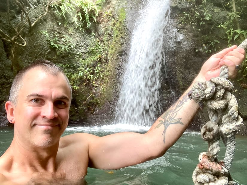 Nick Kembel in a pool of water, facing the camera, holding himself up by holding onto a rope with one hand, with a waterfall spilling into the pool behind him