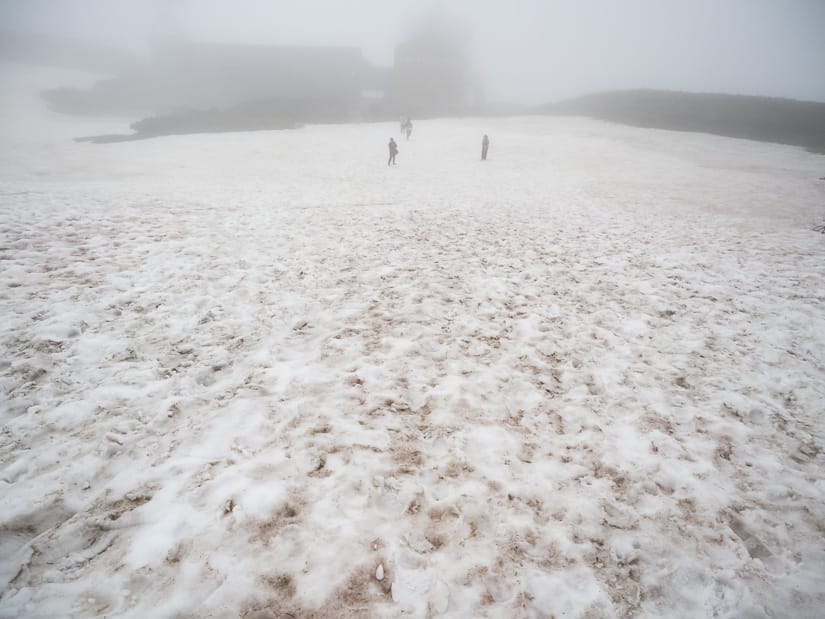 Looking down a snow covered hill, with three figures and a building obscured by mist at the bottom