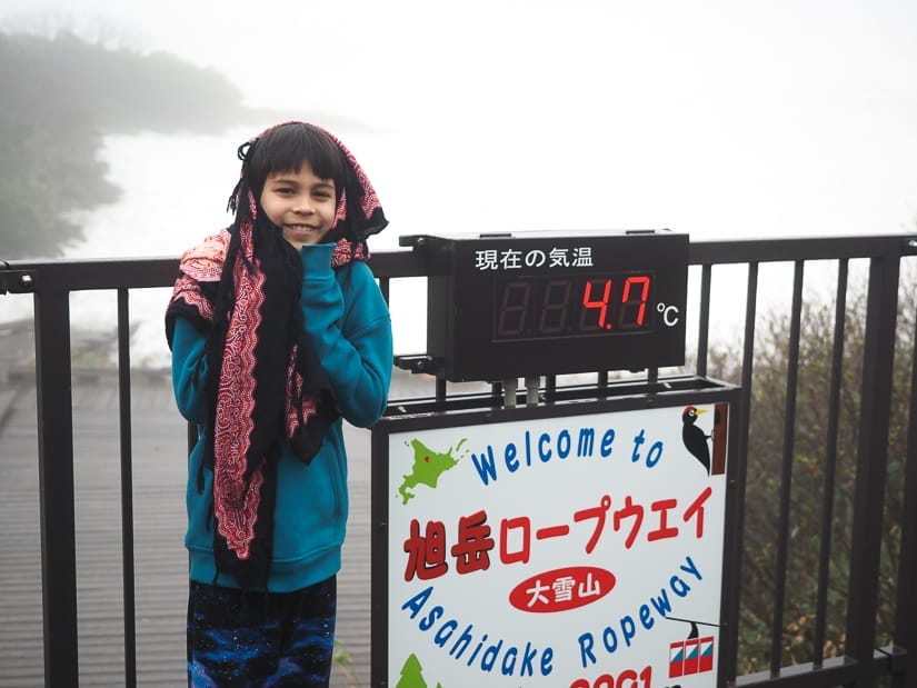Nick Kembel's son Sage standing beside a temperature sign which indicates 4.7 degrees C and a sign that says "welcome to Asahi-dake ropeway), with a snowy scene behind him