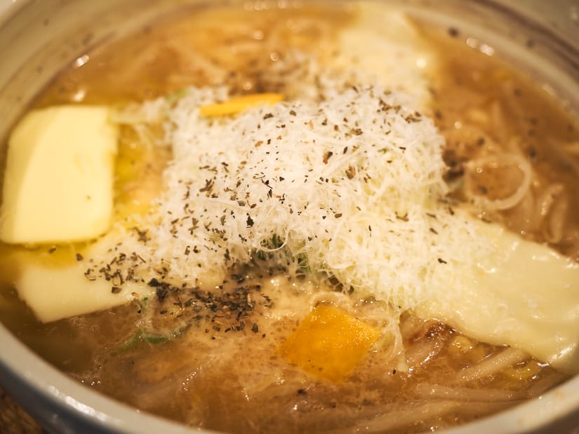 Close up of a bowl of ramen with different types of cheese on top and two chunks of butter in it