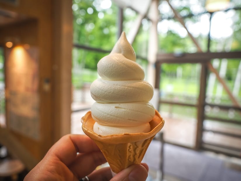 A hand holding up a cone with slightly yellowish soft serve and some windows and greenery outside in the background