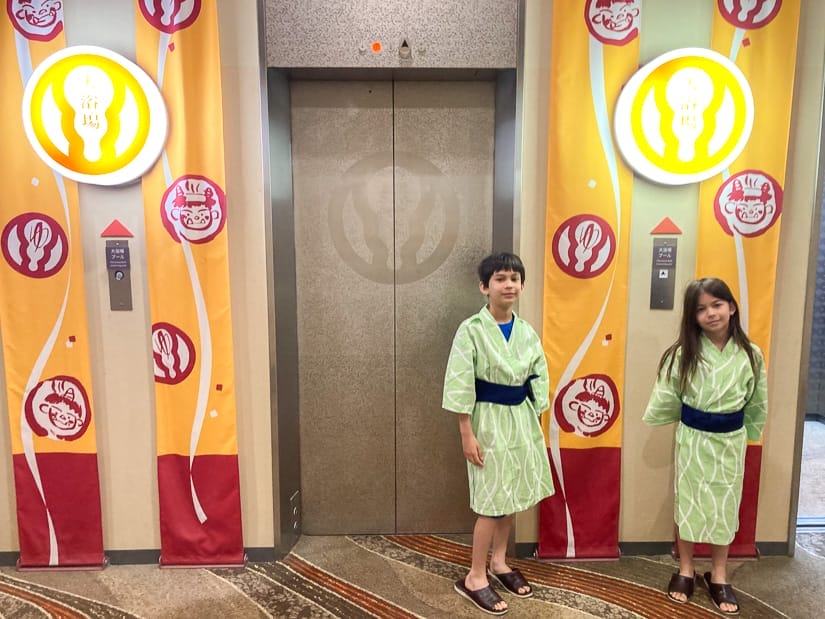 Two kids wearing green traditional Japanese robes and standing in front of an elevator in an onsen resort