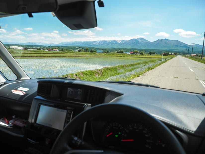 A shot from the angle of a person driving a car on the left side of the road and right side of the car, with rice paddies beside the road outside and mountains in the distance