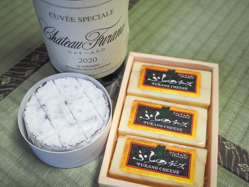 A round white cheese and box of three rectangular cheeses which say "Furano Cheese" on them, plus a bottle of Furano wine, on on a Japanese tatami mat
