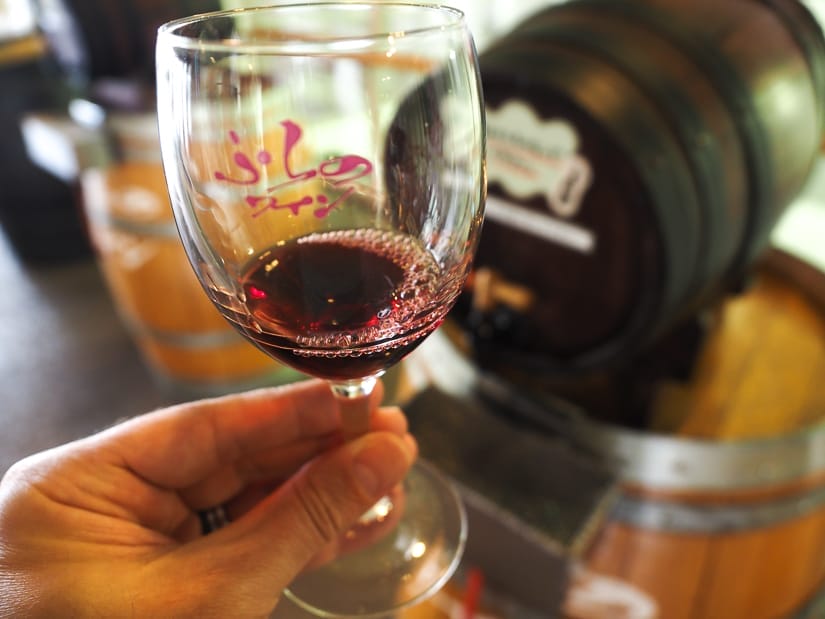 A hand holding up a wine glass with a small amount of red wine in it, with a blurred wine barrel behind it