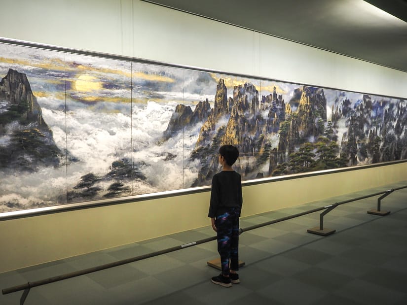 A young boy shot from behind as he looks at a very long painting on a wall in front of him of a natural scene of steep towers of rock surrounded by clouds