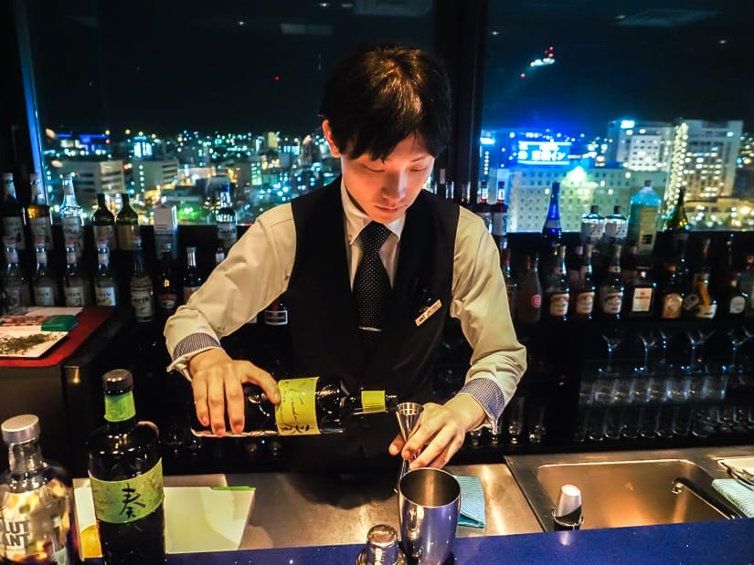 A Japanese bar tender in suit making a cocktail at the bar with the scene of a city at night behind him
