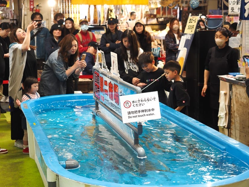 Some people standing around a tank filled with squid, and one kid is fishing for the squid while his mom takes a photo of him