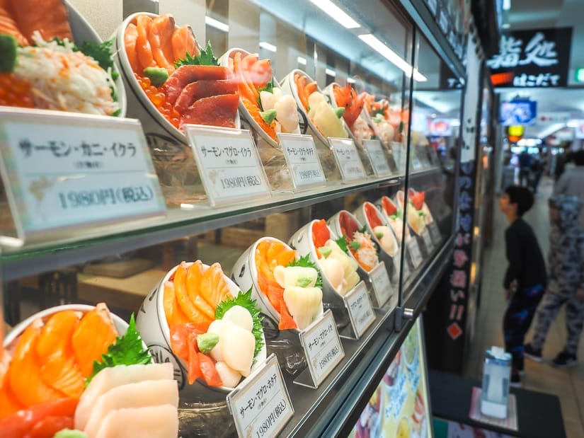 Looking sideways at a glass display with rows of plastic models of seafood dishes with some people visible down the hall on the right