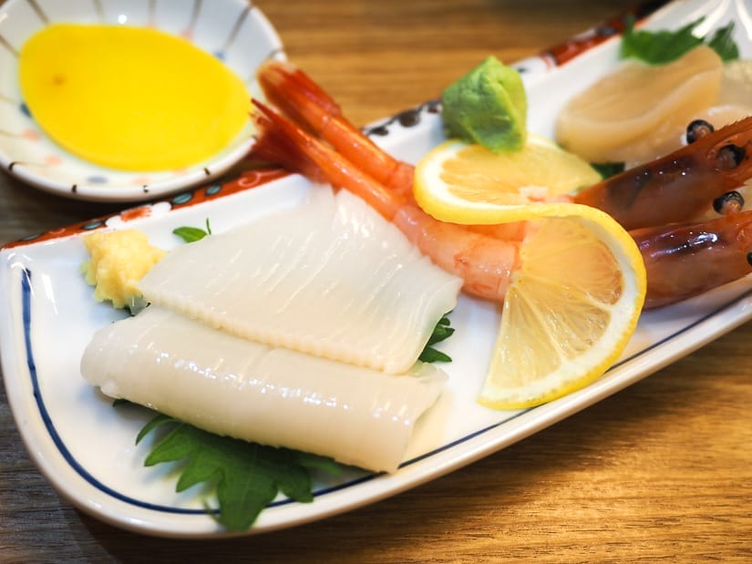 A long white plate with strips of raw squid on it, some lemon slices, and a giant shrimp