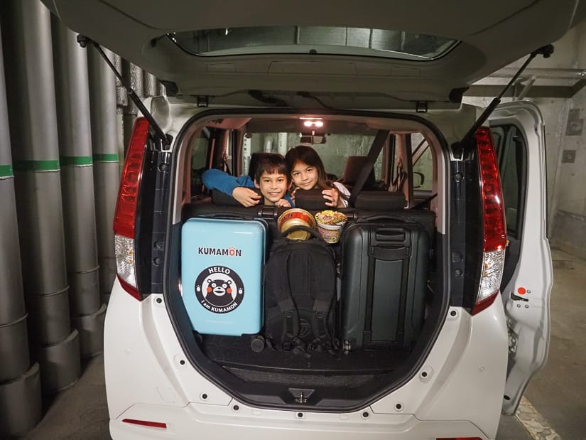Looking into the open trunk of a white car from behind, which is stuffed with luggage, and two kids are sitting in the back seat and looking back out towards the camera