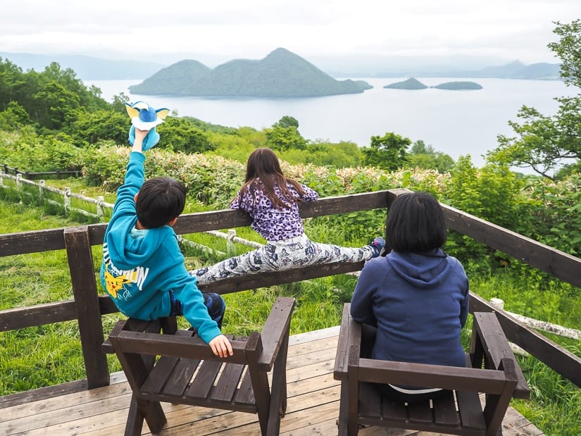 A woman and her two kids shot from behind on a lookout platform, the woman sitting in wooden chair, son sitting on arm of chair and holding up a pokemon stiffie, and daughter is doing the splits on a weekden railing, while they are looking out at a view of a volcanic crater lake