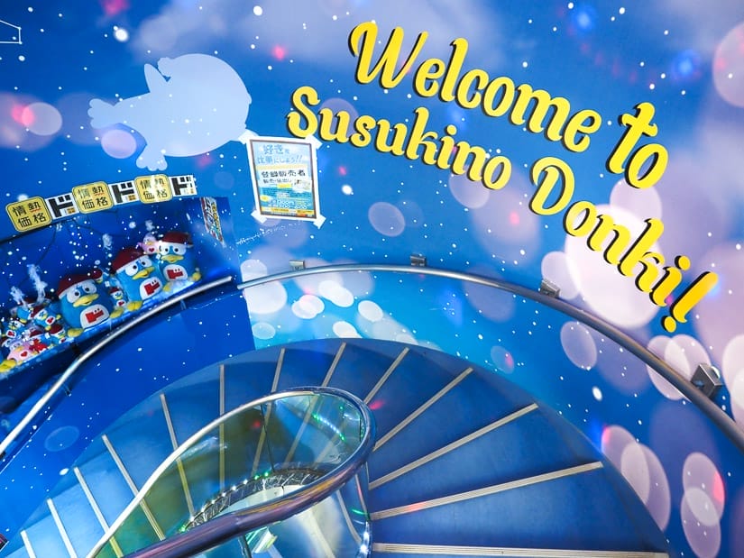 Looking down a curving staircase painted blue, with the wall painted in blues and purples and the words "welcome to Susujino Donki" in yellow on the wall