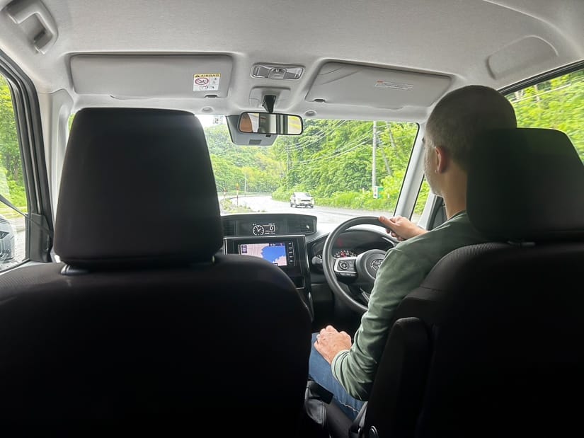 Nick Kembel driving a car while sitting on the right side of it, shot from the back seat, with forest beside the highway out the front window