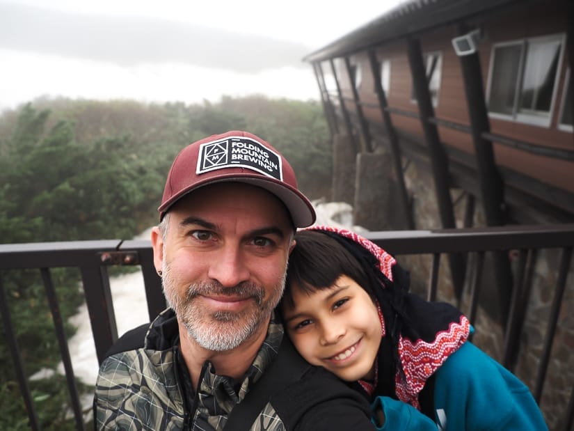 Nick Kembel and his son Sage posing for the camera, with Sage's head on his dad's shoulder, and a cabin with snow beside it behind them