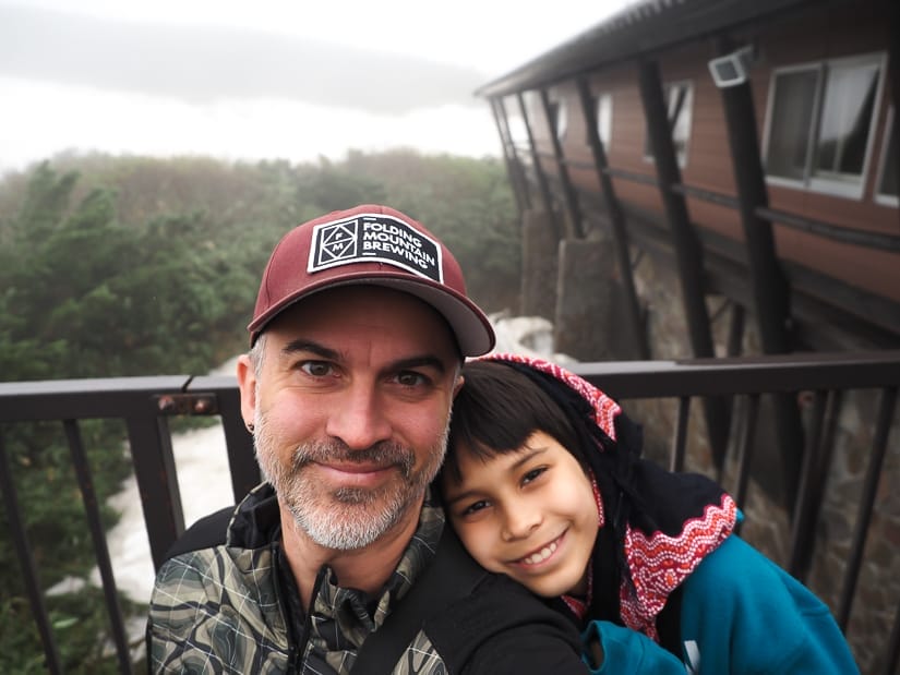 Nick Kembel and his son Sage posing for the camera, with Sage's head on his dad's shoulder, and a cabin with snow beside it behind them