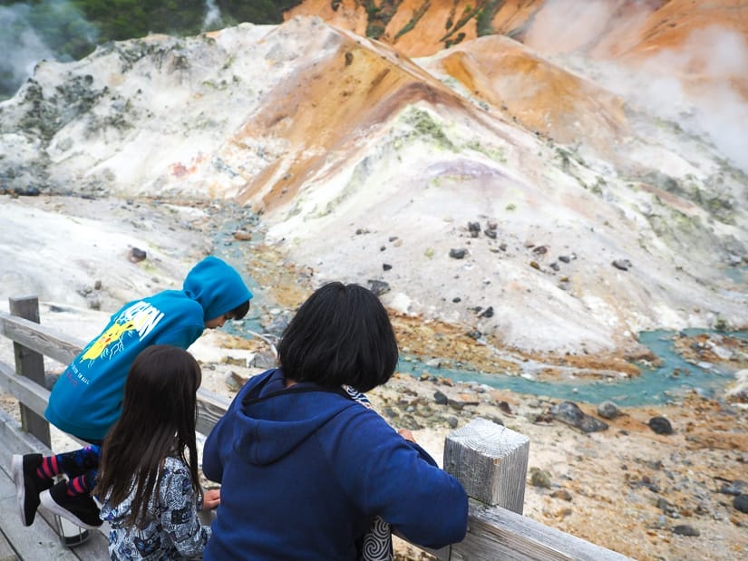 A mother and two kids shot from behind as they look down over a wooden railing at a blue creek running through a steaming gray and orange volcanic crater