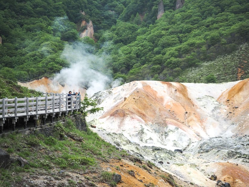 Two kids standing on an elevated walking path in the distance, raising their arms in the air, with a steaming volcanic crater below them