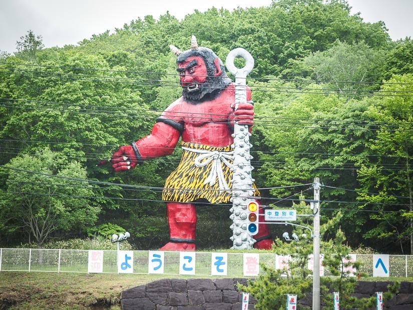 A large red demon statue holding a big spiky stick on the side of a road