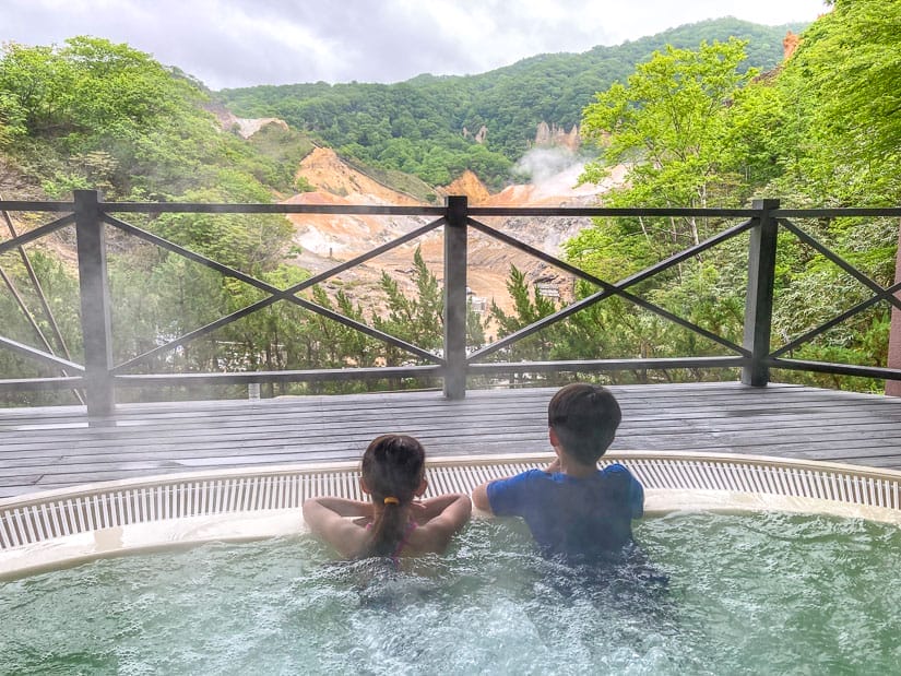 Two kids in a hot tub, shot from behind as the look out at a view of a volcanic caldera