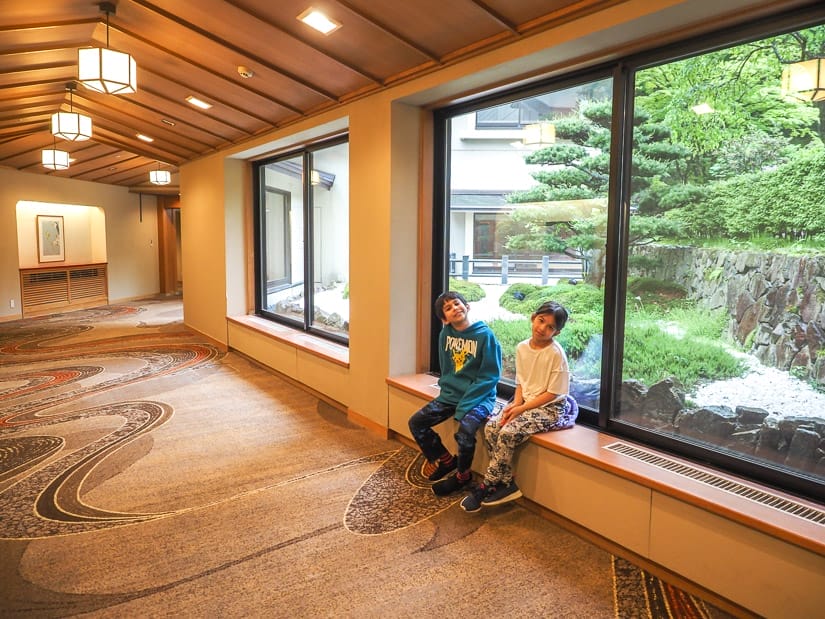 Two kids sitting on a window frame in a hotel hallway, with a Japanese garden outside the window