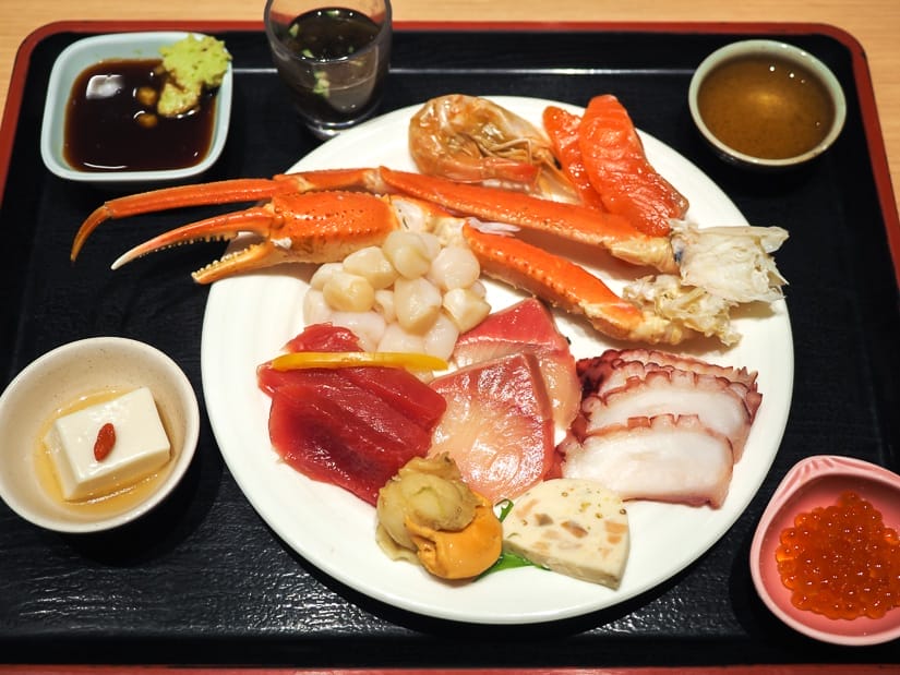 A white plate filled with seafood including large crab leg, sashimi, shrimp, scallops, on top of a black tray, which also contains small dishes of tofu, soy sauce and wasabi, and fish roe