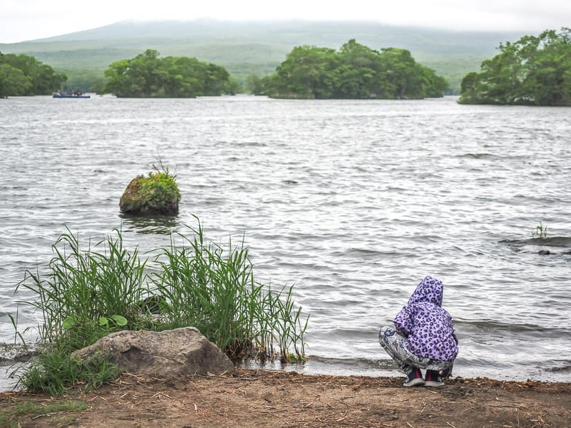 A young girl wearing purple jacket shot from behind as she kneels down on a small beach, with a lake in front of her with rough water and cloudy sky obscuring a volcano on the other side of the lake