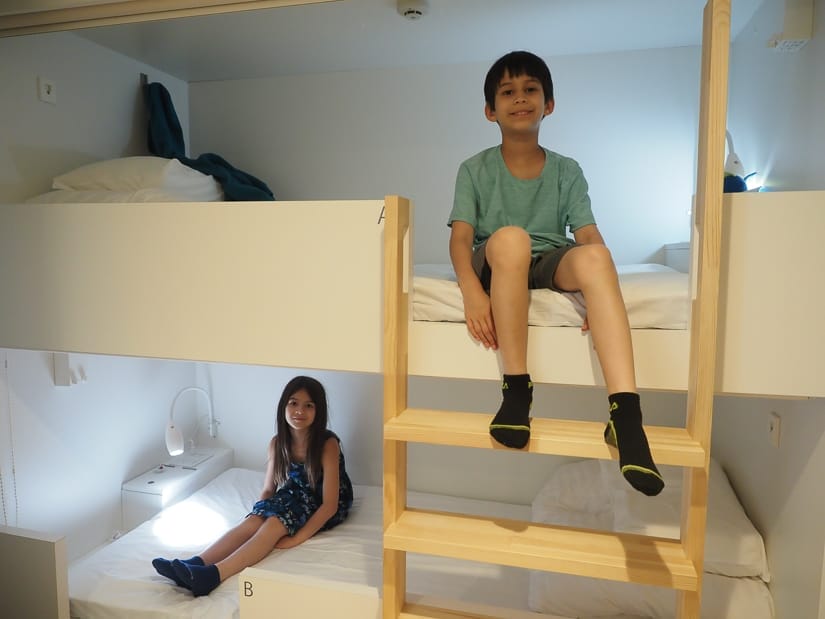 Two kids in a hostel bunk bed, the boy sitting at the top of the ladder on the top bunk and the girl sitting beside the lit lamp on the bottom bunk