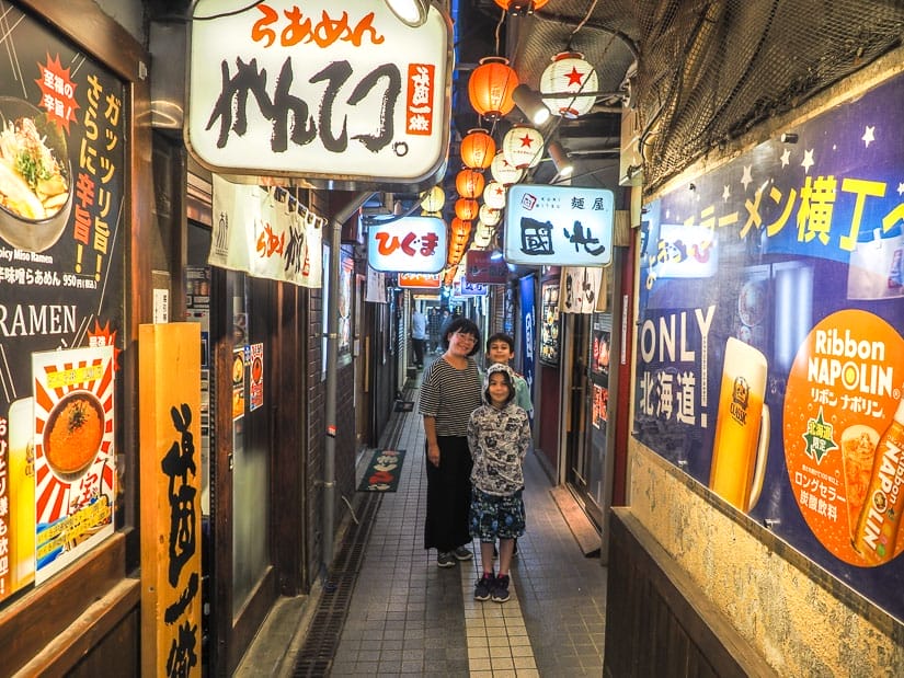 A mother and two kids standing in a narrow alley with lit up ramen signs on either side and red lanterns hanging above