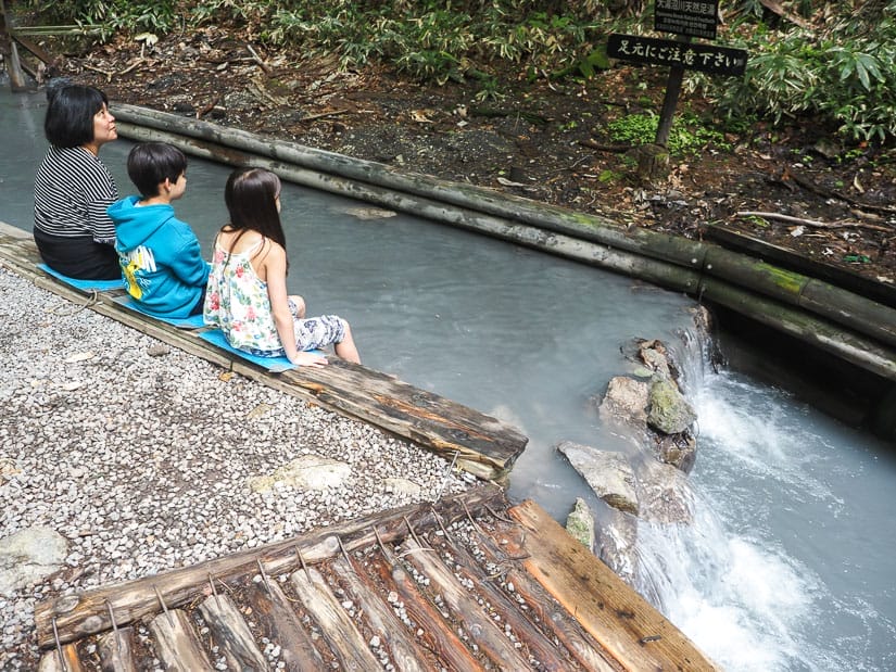 A mother and two kids sitting on a wooden beam on the side of a hot water creek soaking their feet in it