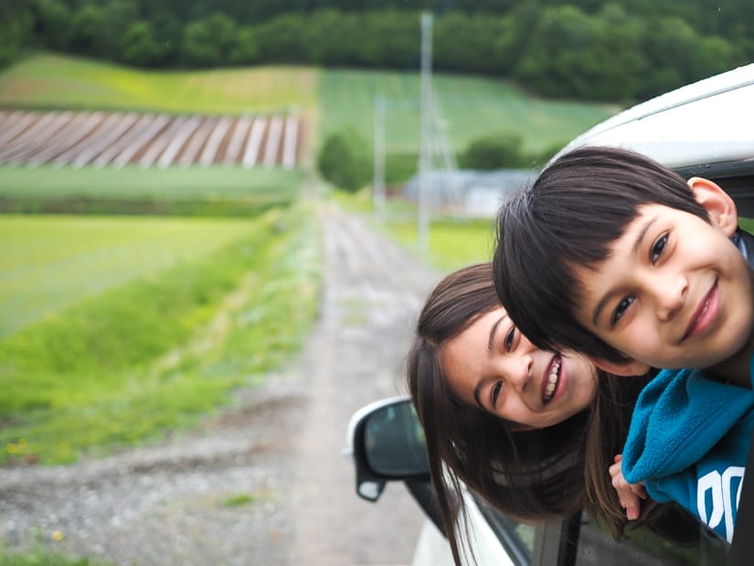Two kids looking out and backwards from the window of a white car which is parked on a small road beside some farm fields