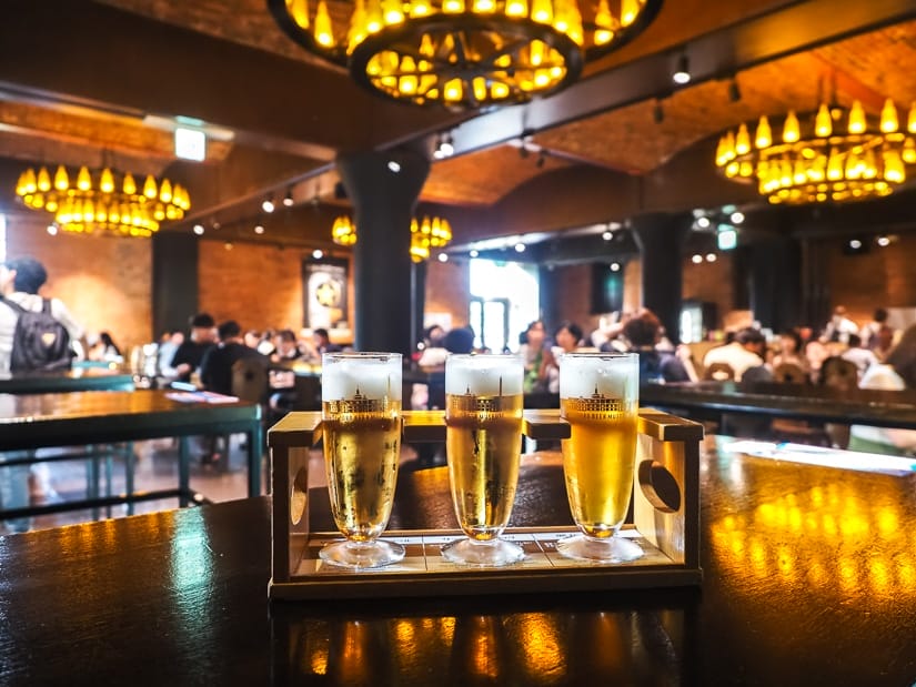 A rack of three beer samples on a table, with a beer hall behind included round chandeliers and people sitting at tables drinking beer