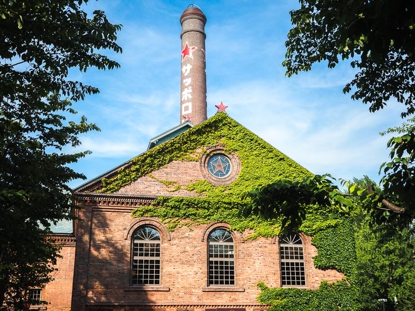 Exterior of an old red brick building with green foliage growing on the sides, red smoke stack with symbol of Sapporo beer on it, and all frame by some tree branches