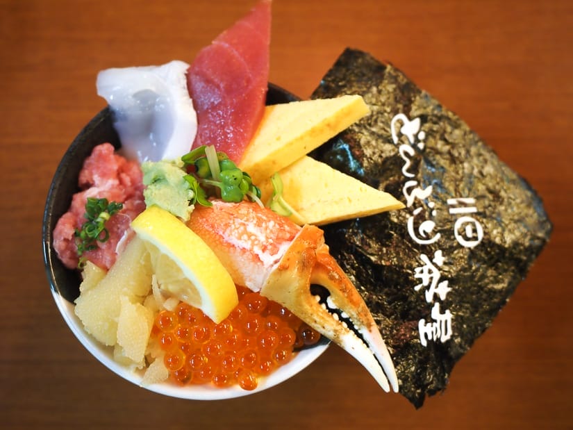 Close up of a bowl of seafood don, with egg, crab claw, fish eggs and various sashimi on rice, with a large piece of seaweed sticking out and some Japanese characters written in white on it