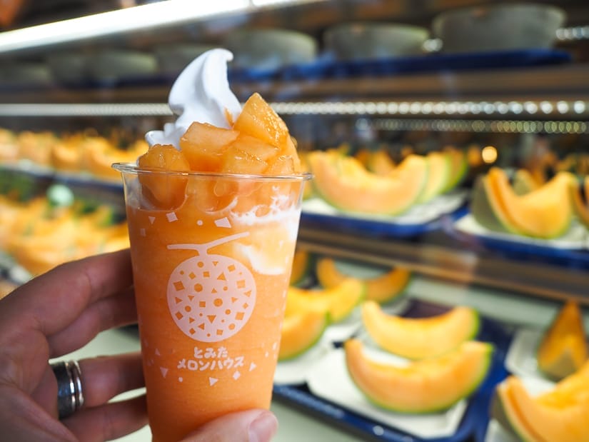 A hand holding up an orange melon smoothie with melon chunks and white soft serve ice cream at the top, in front of a glass display full of slices of melon for sale
