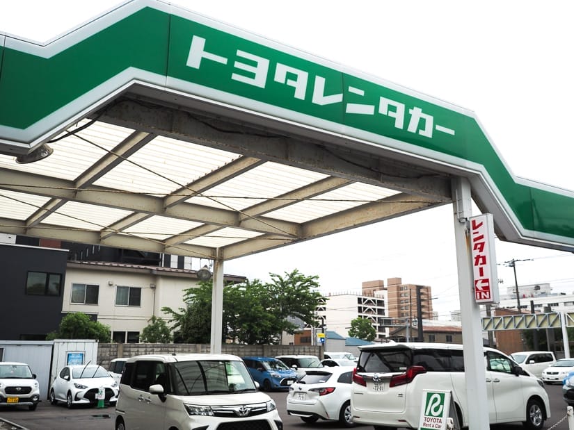A Toyoto car rental lot with the name in white Japanese writing on a green sign above and some cars in a lot below waiting to be rented