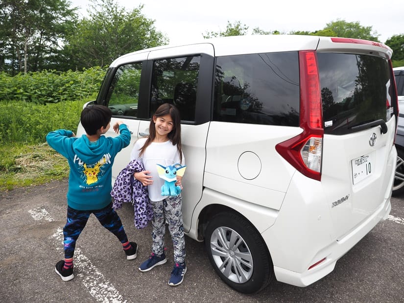 Two kids making silly poses in beside a white cube-shaped Toyota Roomy car in a parking lot
