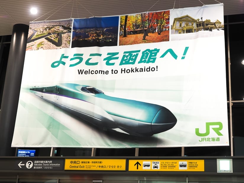 A large sign hanging in a train station which shows a Japanese bullet train, scenes of Hakodate, and says "welcome to Hokkaido"