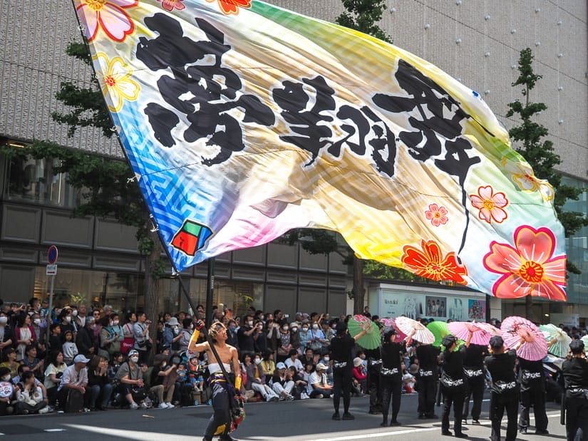 A Japanese man in costume with no shirt waving a huge colorful flag with Japanese characters on it, with some other performers to the right holding up colorful umbrellas and some spectators standing on the street watching