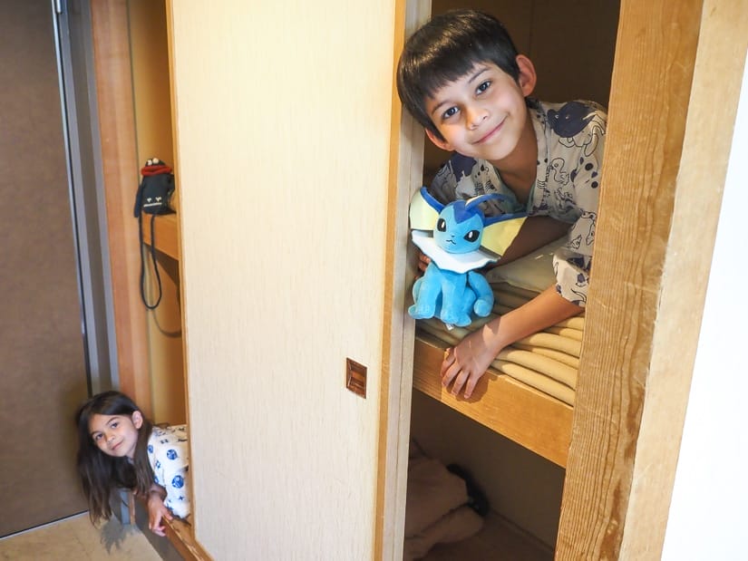 Two kids peering out from a two-level closet, the boy is on the top and peering out from the right side, holding out a blue stuffie, while the girl is on the bottom, peering out from the left side