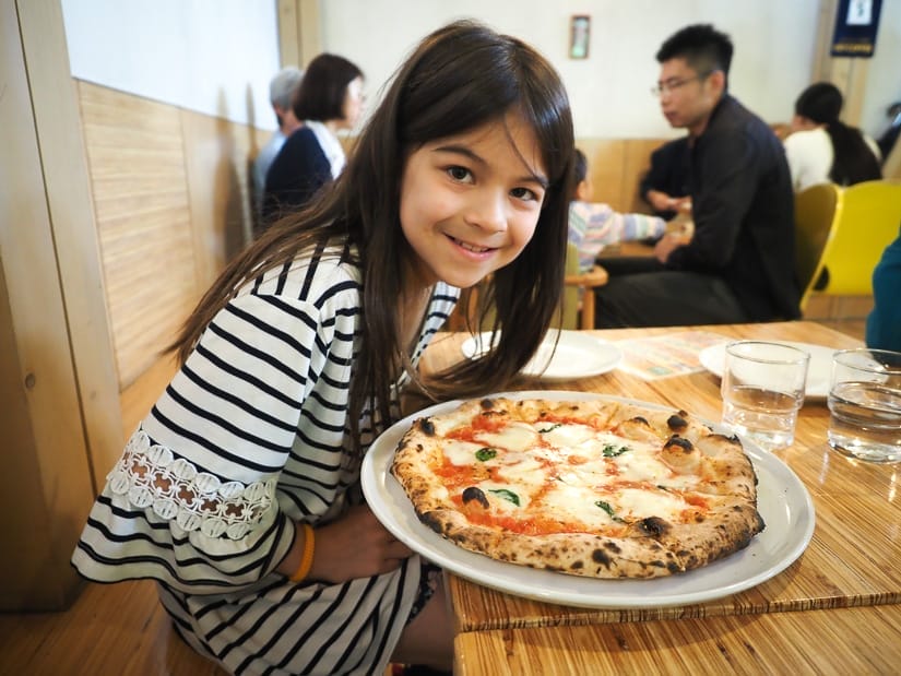 A young girl where a large black and white striped shirt sitting at a table, smiling at the camera, with a large cheese pizza on the table in front of her
