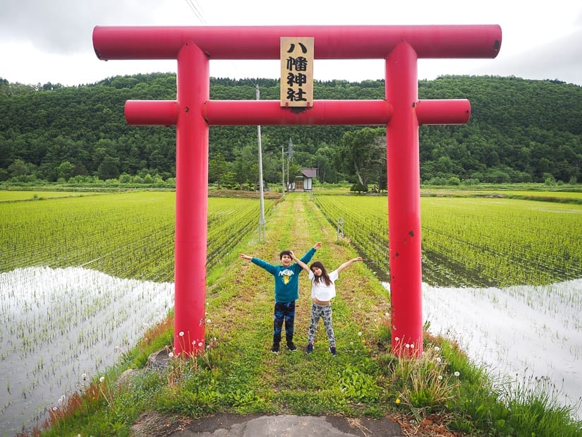 Two kids raising their arms while standing below a red torri Japanese gate with rice paddies on either side of them