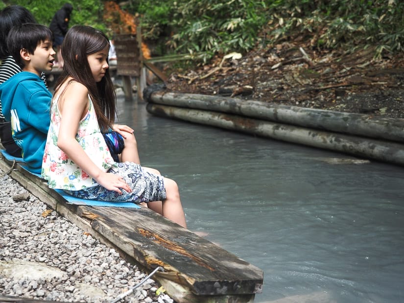 Looking up a small creek with two kids sitting on wooden bench on the side, soaking their feet in the water