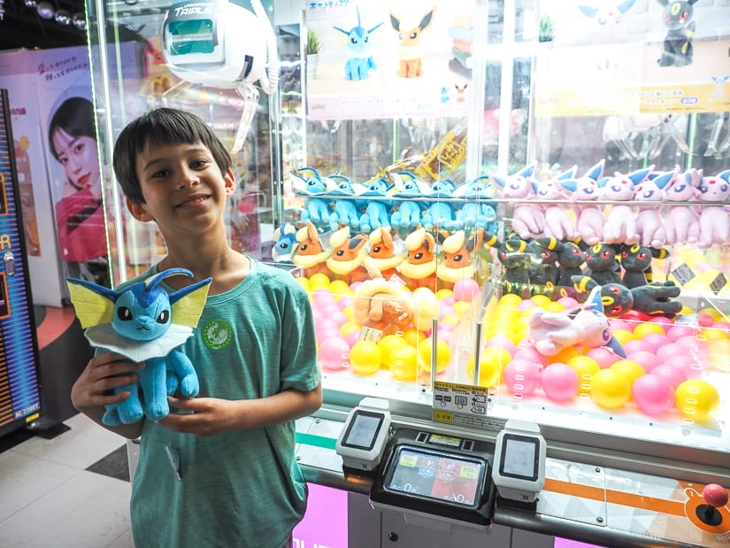 A smiling boy holding a pokemon stuffie that he just won from the claw machine behind him