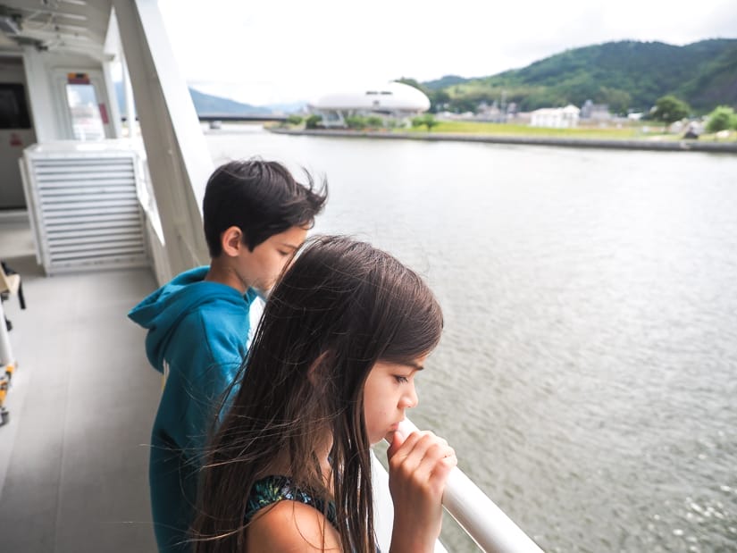 Two kids on a ferry, looking over the side, as it sails down a river, with a museum visible on the riverbank in the distance