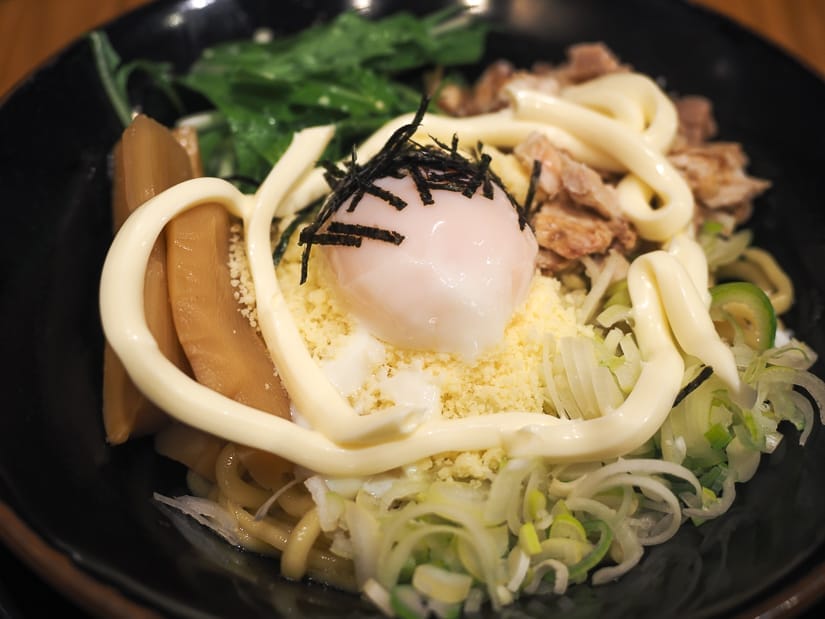 Close up of a black bowl of ramen topped with powdered cheese, soft boiled egg, mayonnaise, bamboo, seaweed flakes, and green onions