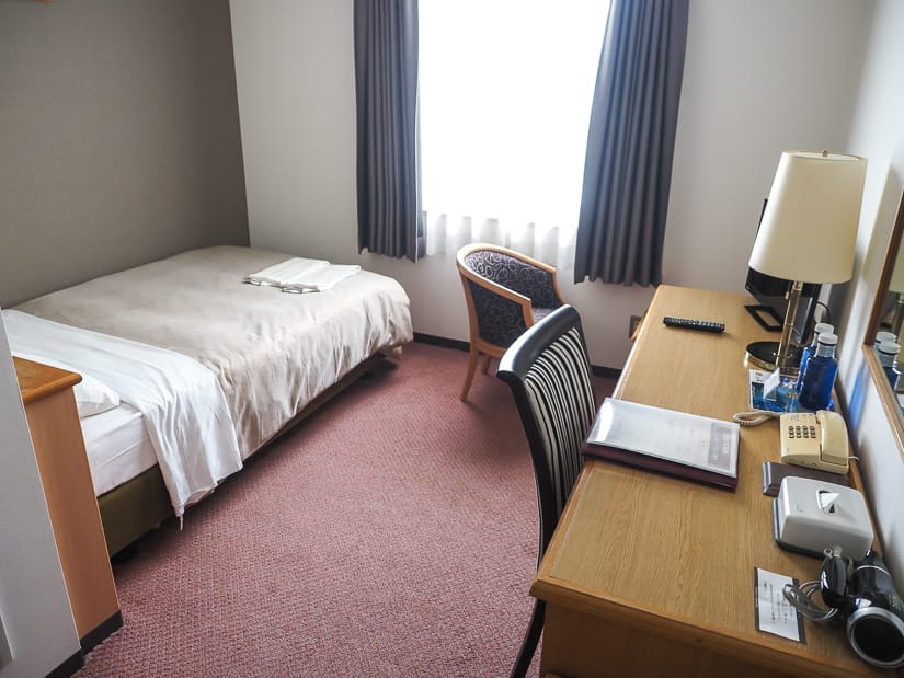 Looking inside a hotel room with small bed on left side and desk, chair, phone, etc on right side