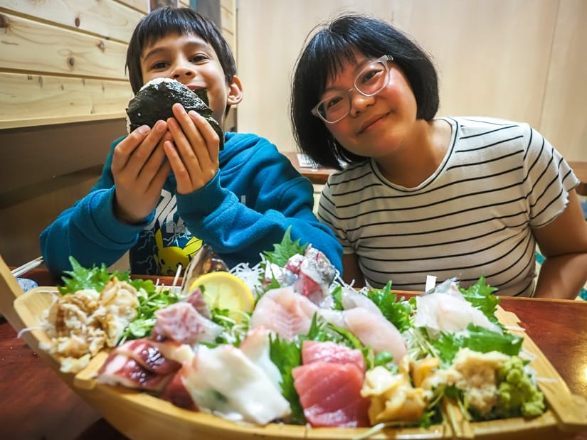 Nick Kembel's wife and son at a table in a restaurant facing the camera, with a boat of sashimi on the table in front of them, and the son is holding up an onigiri wrapped in seaweed