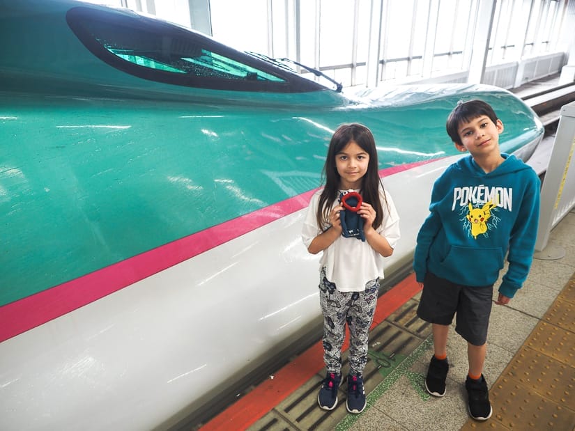 A young boy and girl posing beside the front car of a turquoise shinkansen bullet train, and the girl is holding up a stuffie