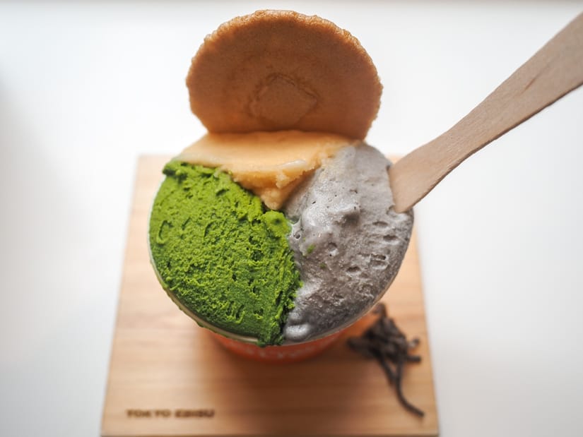 A small paper bowl of ice cream with three kinds (matcha, sesame, brown rice) on a little square wooden platter, with some tea leaves on it, wound cookie on top, and a wooden spoon sticking out of it